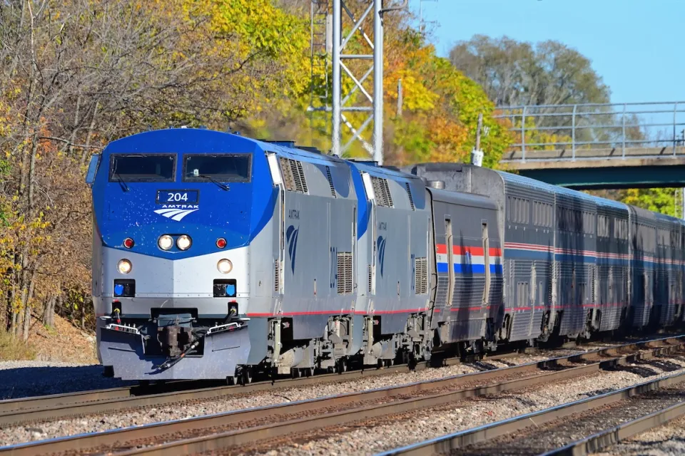 Blue and silver Amtrak train with vibrant green and yellow foliage in background.